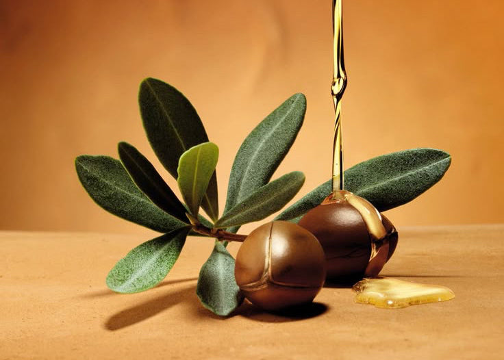 Olive oil being drizzled over olives with leaves on a brown background